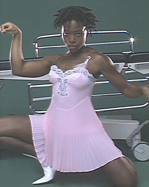 Athletic Black woman in pink slip dress posing in front of medical equipment. She has short dreadlocks and wears white boots.