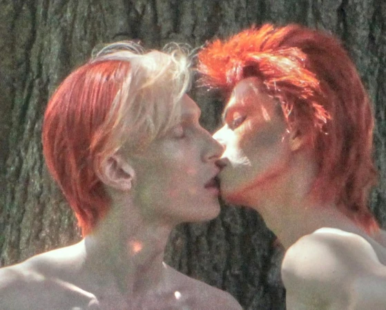 Two people with dyed red hair kissing outdoors in front of a tree trunk, captured in a grainy, lo-fi aesthetic by Sharna Osborne.