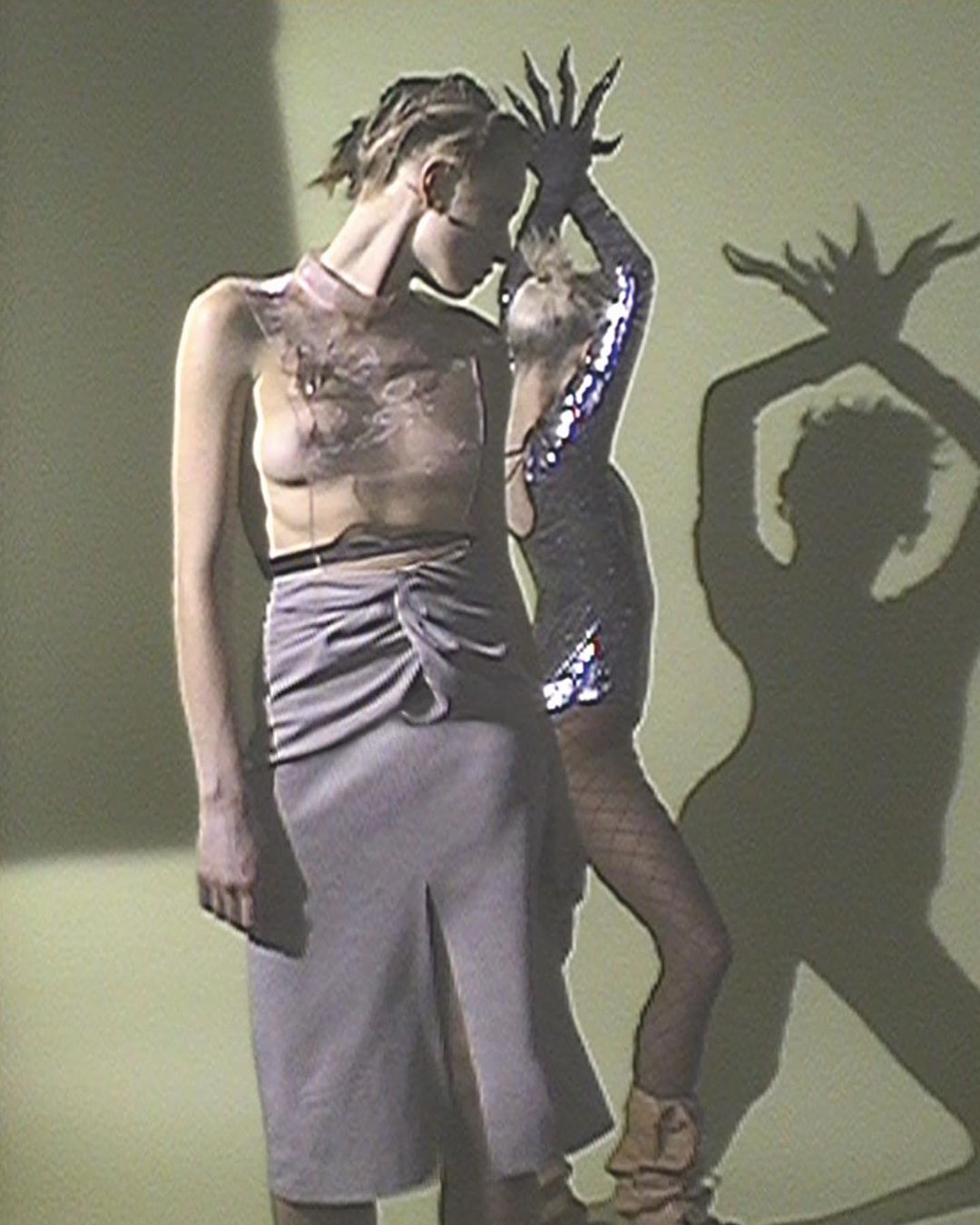 Two women in fashion-forward outfits stand in front of a lime green wall, with one woman casting a shadow puppet with her hands.