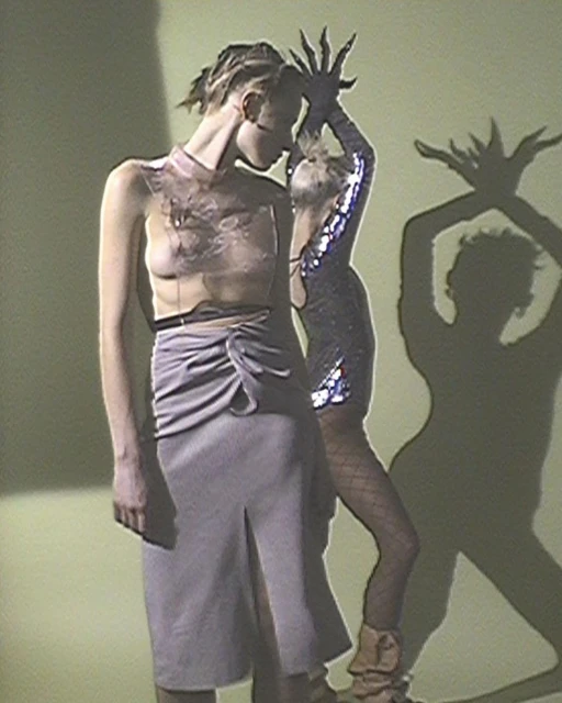 Two women in fashion-forward outfits stand in front of a lime green wall, with one woman casting a shadow puppet with her hands.