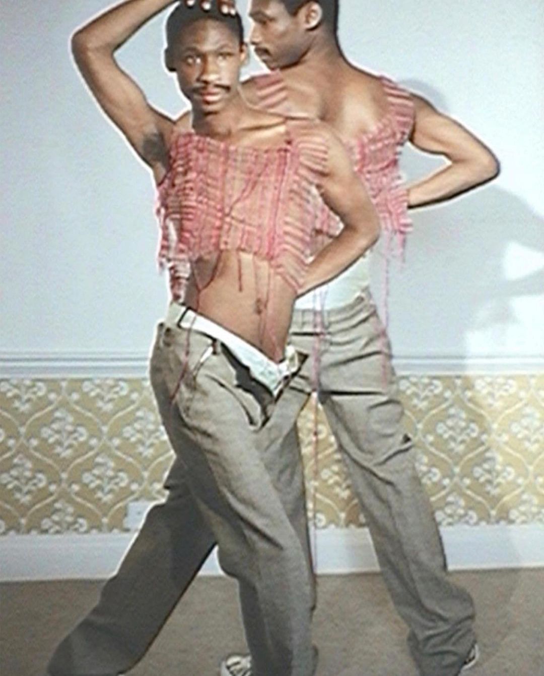 Two young Black men pose indoors wearing tops made from string. The subjects are wearing slacks with decorative patterned walls in the background.