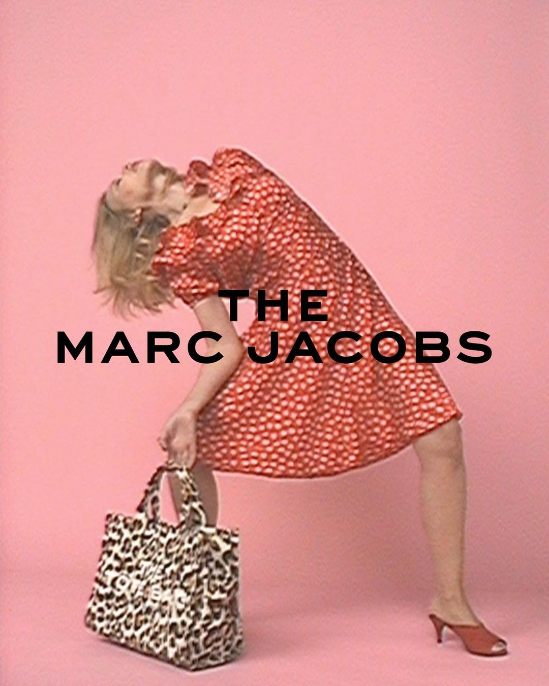 A slender woman in a red patterned dress poses with a Marc Jacobs leopard-print tote bag against a solid pink backdrop.