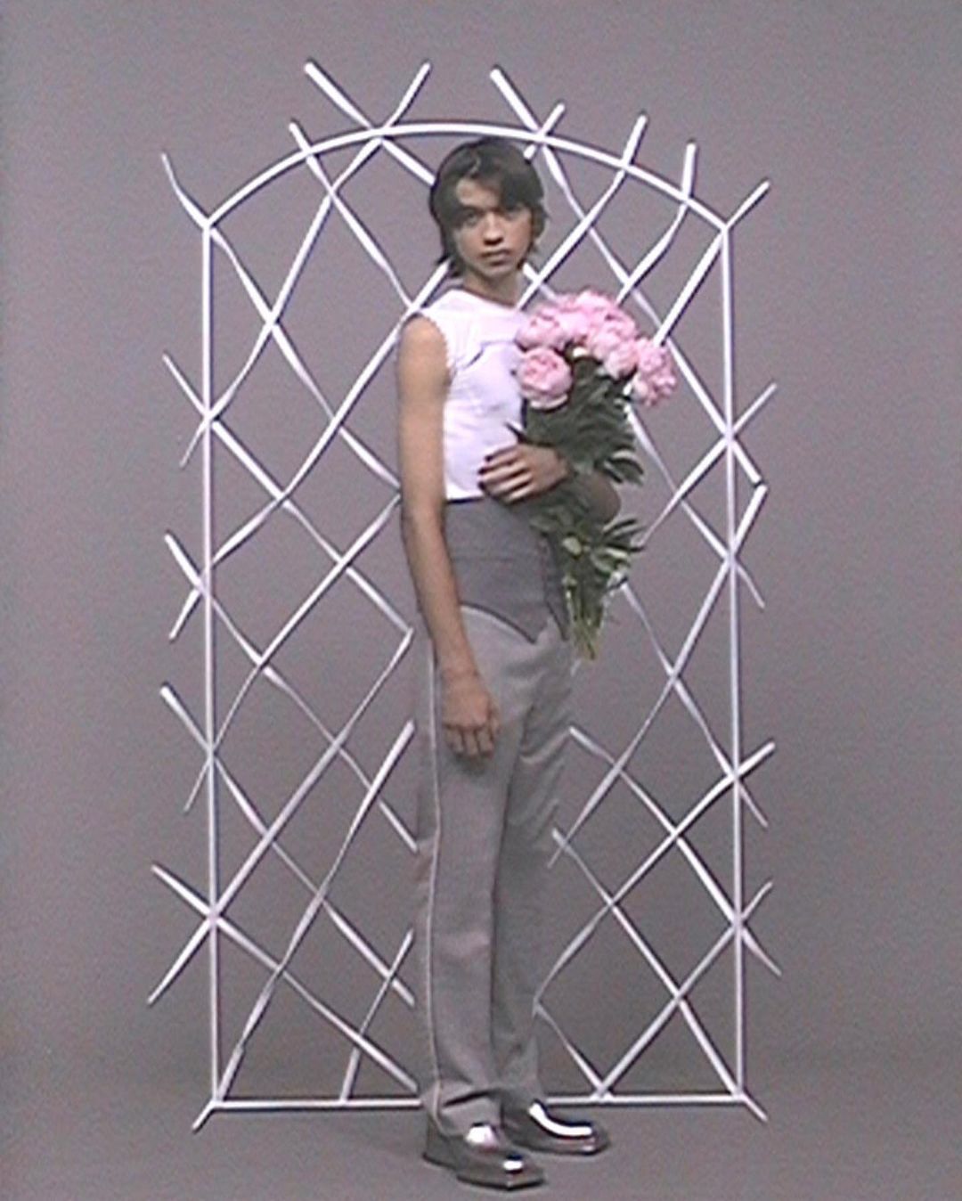 Full-length studio shot of androgynous person holding pink peonies with a white ribbon structure and gray backdrop. Style and fashion.