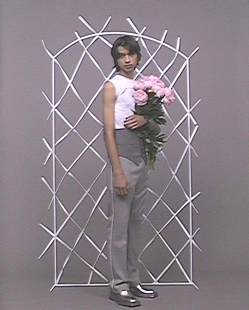 Full-length studio shot of androgynous person holding pink peonies with a white ribbon structure and gray backdrop. Style and fashion.