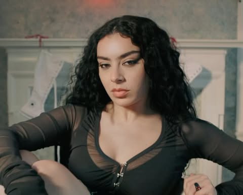 Close-up of a woman with long, dark curly hair, wearing a black mesh top, gazing at the camera with a serious expression in an indoor setting.