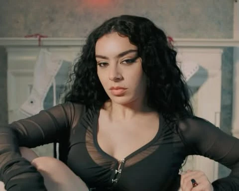 Close-up of a woman with long, dark curly hair, wearing a black mesh top, gazing at the camera with a serious expression in an indoor setting.
