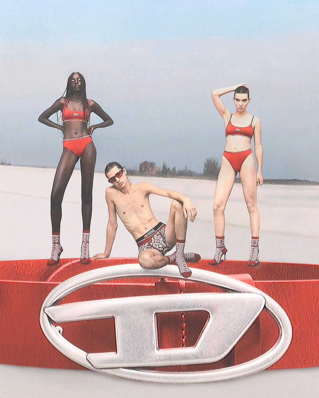 Fashion shot: three models in swimwear and high heels posing on a sand dune with a Diesel belt in the foreground, advertising fashion and style.