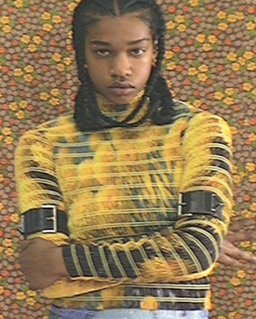 Close-up portrait of a person with braided hair wearing a yellow patterned turtleneck top against a floral wallpaper background.
