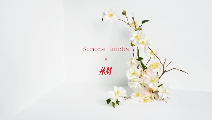 White camellias and bare branches create a border with Simone Rocha x H&M logo on a white seamless backdrop in a studio setting.