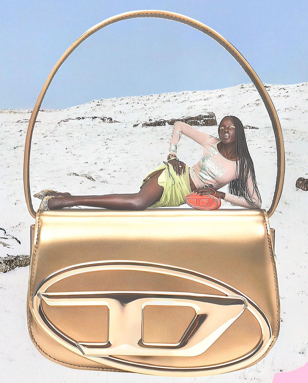 Model lying across a giant metallic gold handbag against a snowy landscape background