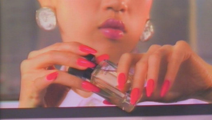 Close-up of hands with long pink nails holding a perfume bottle with reflective earrings and glossy lips in a beauty shot.