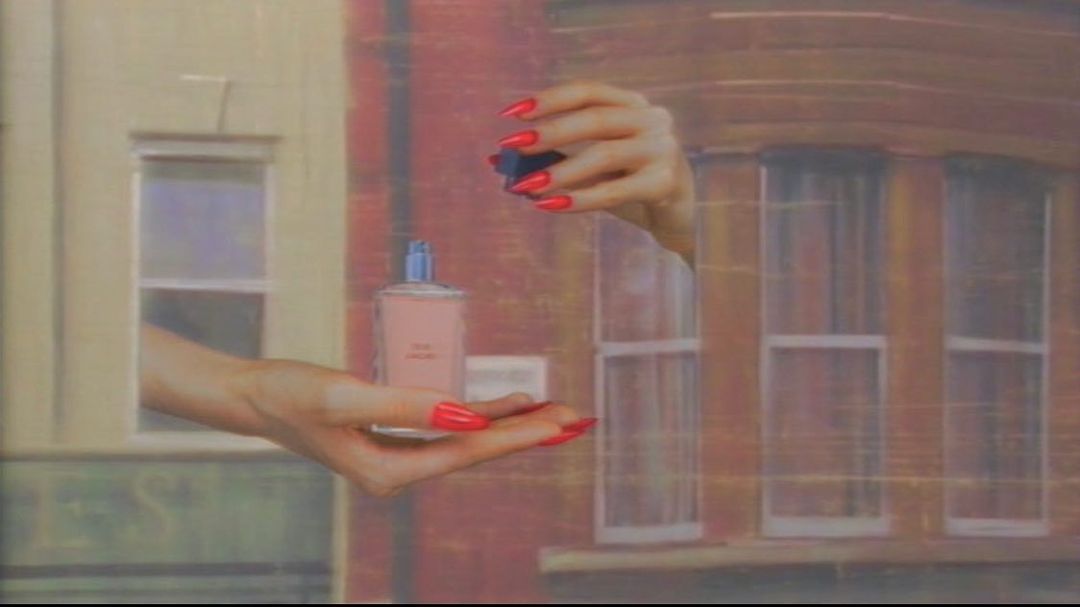 Woman's hands with red nails holding a perfume bottle and its cap against an urban backdrop with visible windows and brick.