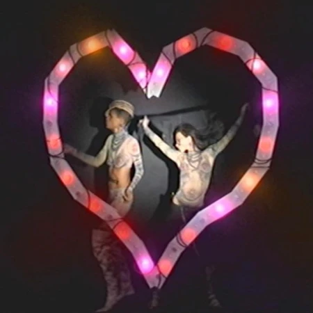 Two people pose within a heart made of illuminated tubing against a black backdrop in a performance setting with creative styling.