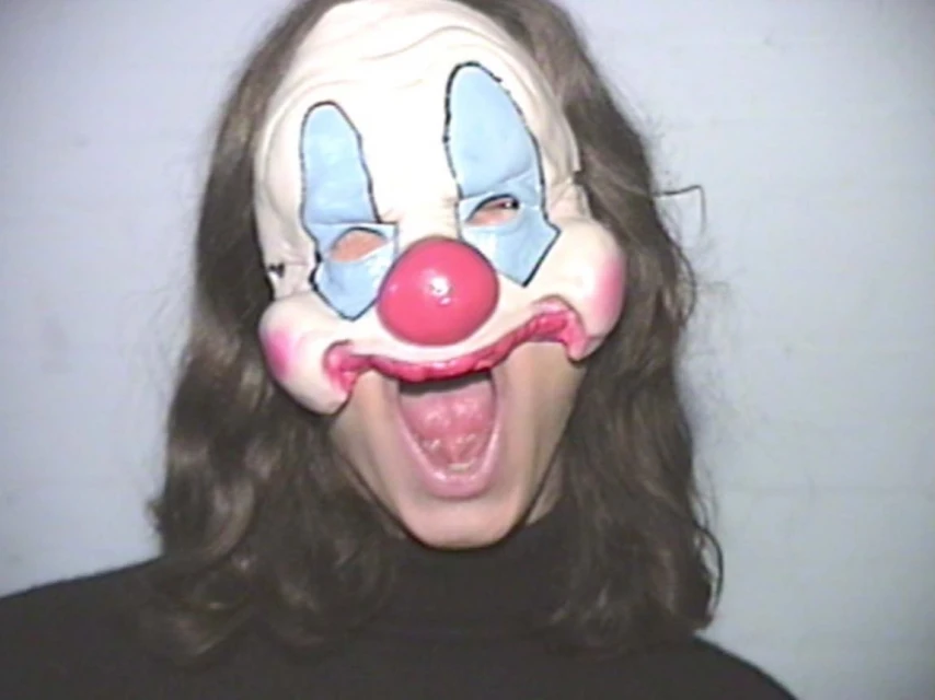 Close-up of a person wearing a clown mask with a pink nose and blue accents, mouth open in a shout, creating a vintage, slightly eerie feel.