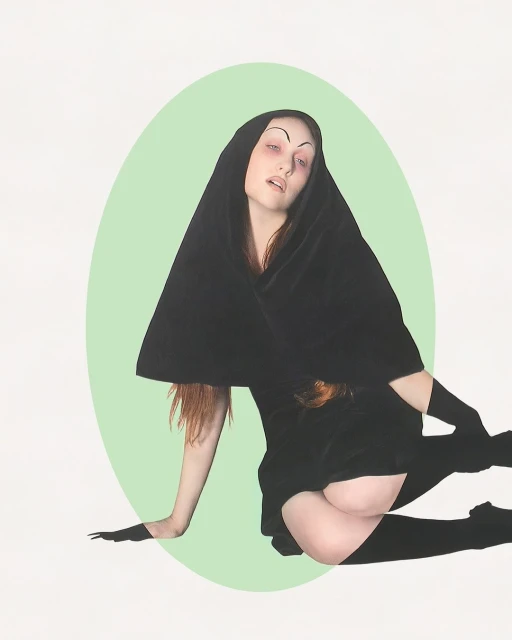Woman in hooded black velvet dress, tights, and gloves posed against pastel green oval and white backdrop.  Artistic fashion portrait.
