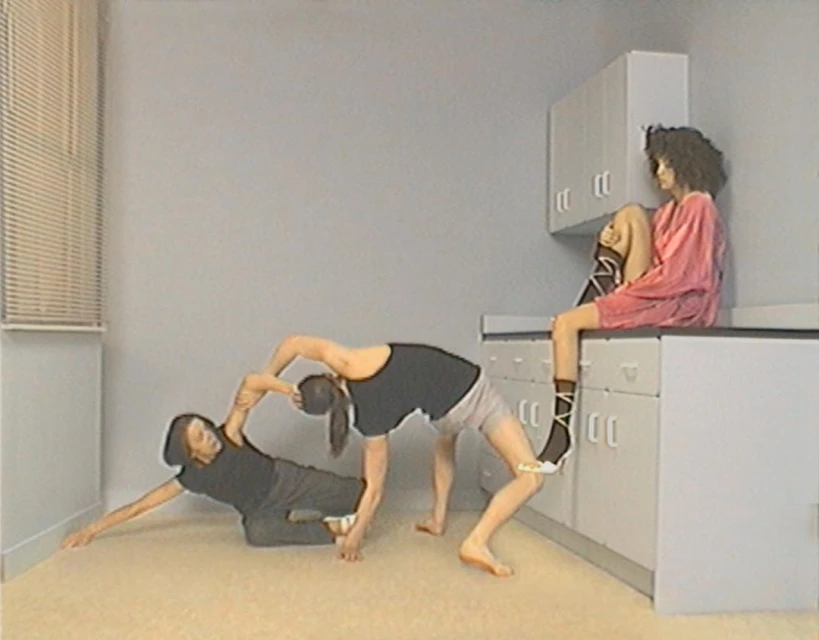 Three figures perform a gymnastic pose in an indoor setting. One figure sits on a counter, and two figures create an arching bridge.