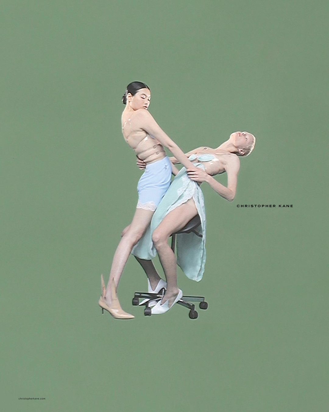Two women in contemporary fashion pose on an office chair against a green background in a studio shot for a fashion editorial.
