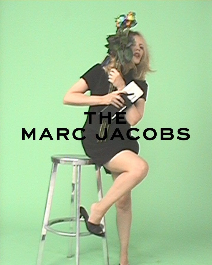Woman in a black dress poses on a stool in front of a green screen, holding flowers and a book with "The Marc Jacobs" text overlay.