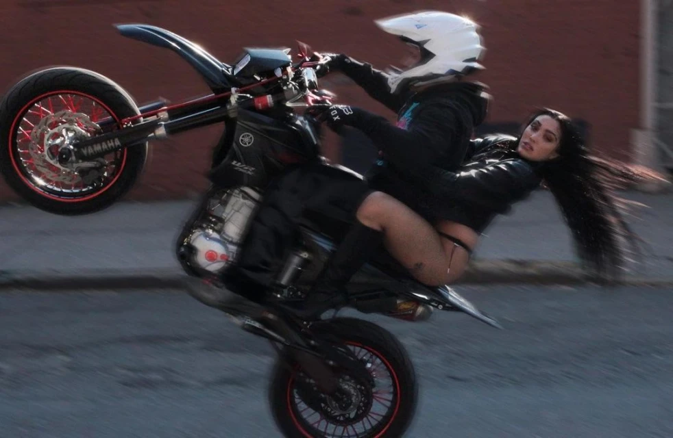 Motorcycle stunt with two women, one riding and one seated on the gas tank wearing black, with a building in the background.