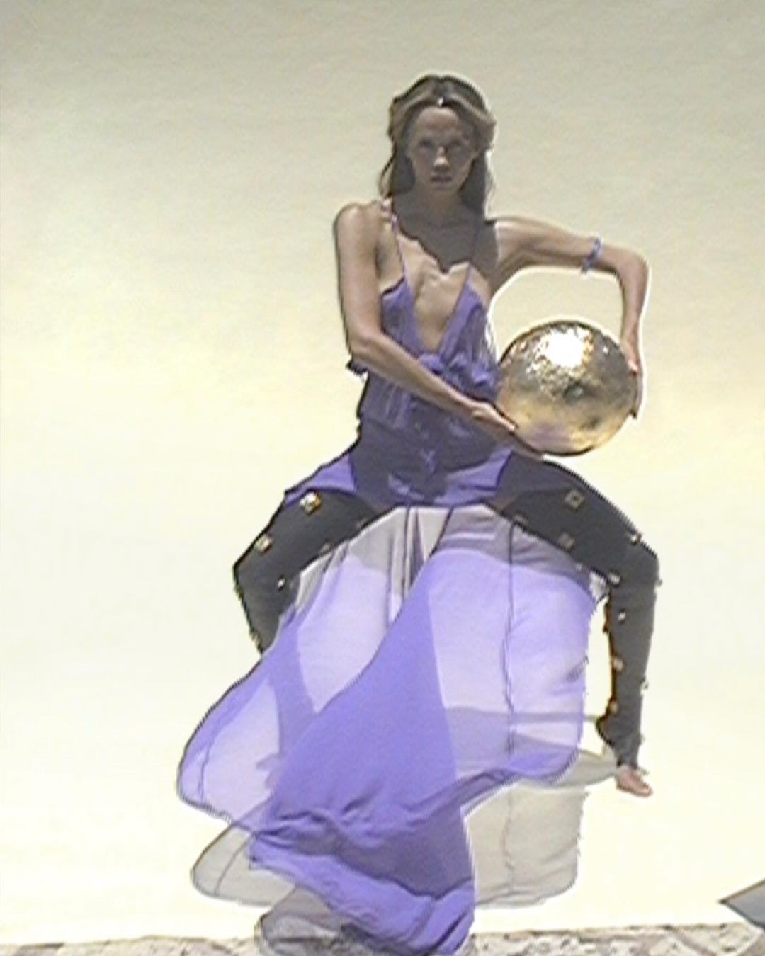 An adult female in a purple outfit poses with a gold ball against a light backdrop, showcasing fashion and artistry in a studio setting.