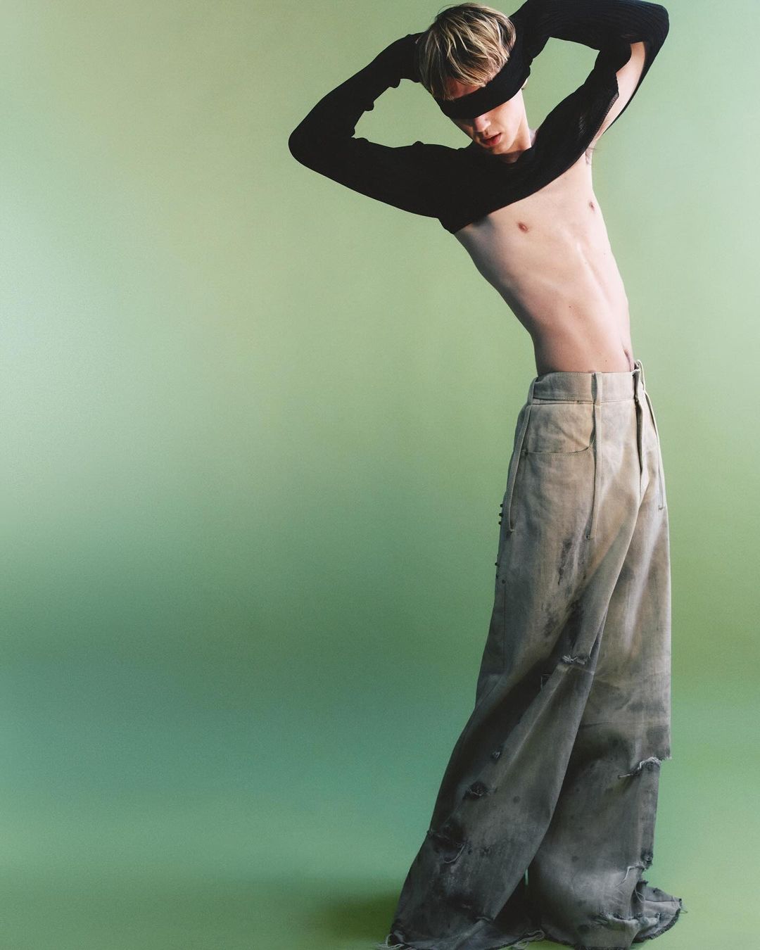 Fashion shot of a blond male model in baggy jeans and a cropped top, posing against a light green background in a studio setting.