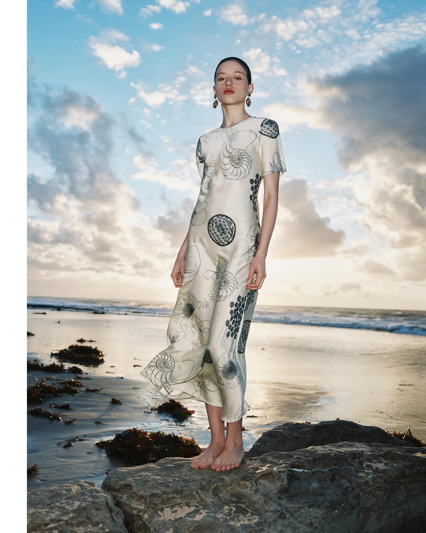 Model standing barefoot on a rocky shore wearing an ivory dress with a shell print against an ocean backdrop.
