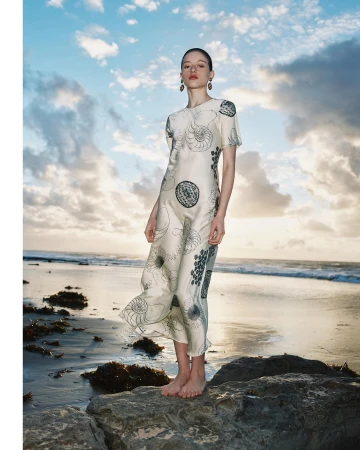 Model standing barefoot on a rocky shore wearing an ivory dress with a shell print against an ocean backdrop.