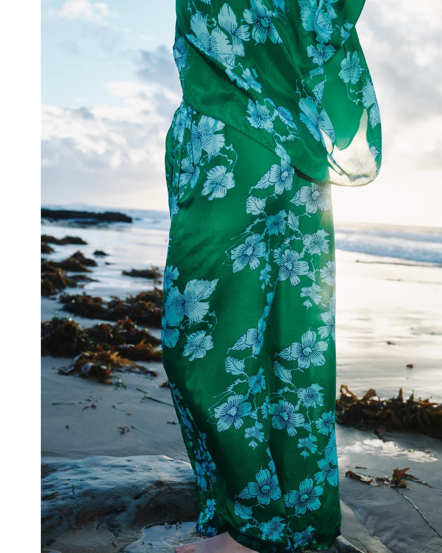 Woman in green floral dress stands on a beach. Ocean waves and sky are visible in the background under soft, diffused sunlight.