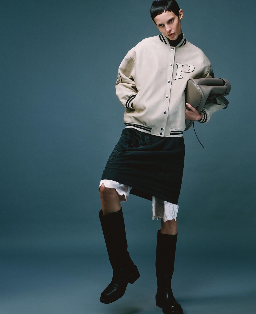 Fashion shot of a model wearing a letterman jacket, skirt, and high boots posing in a studio with a solid blue background.