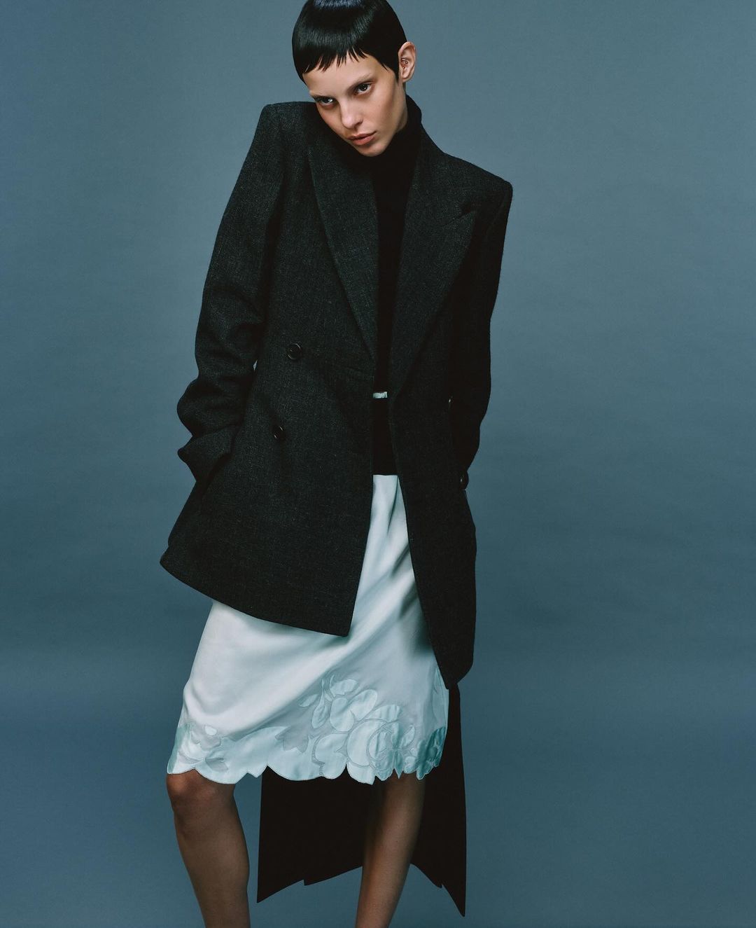 An adult female poses in a dark wool coat and white skirt in a studio, showcasing a stylish fashion ensemble against a neutral backdrop.