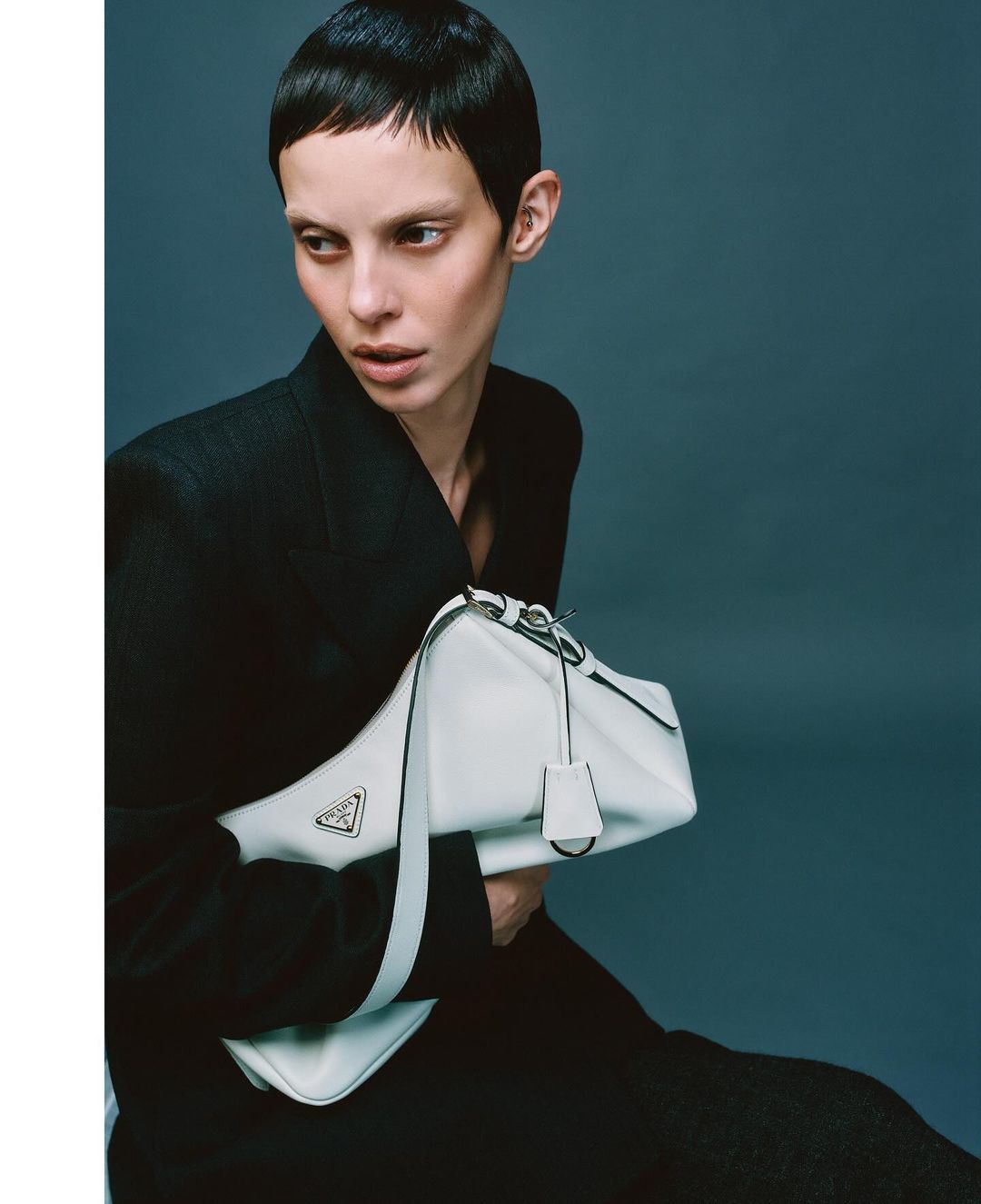Woman with short dark hair poses indoors wearing a black blazer and holding a white Prada handbag in a fashion portrait.