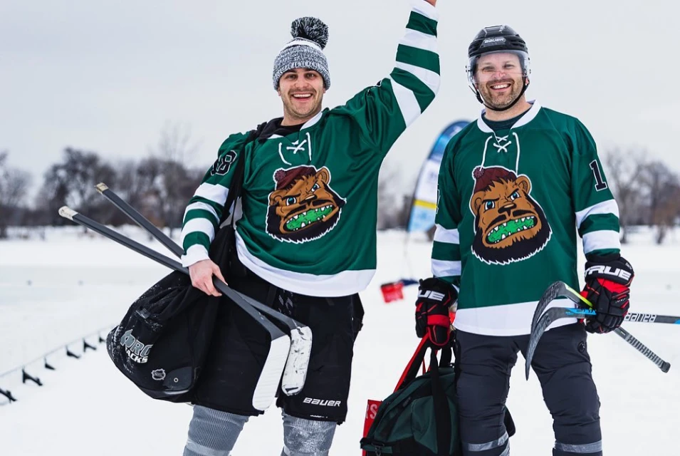 Two smiling men in hockey jerseys stand on a snowy field holding hockey sticks, ready for a game of ice hockey.