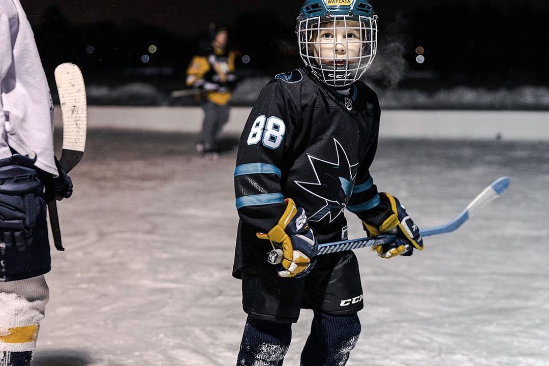 Young hockey player in full gear with stick on an outdoor rink at night. Focus on child in black uniform with team logo.