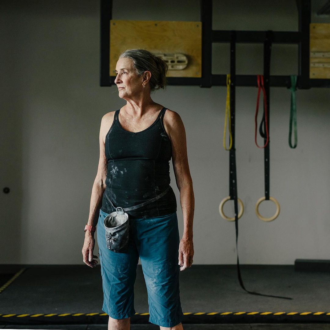 Older woman with chalk on her clothing and a chalk bag, stands in an indoor climbing gym with climbing rings and equipment.