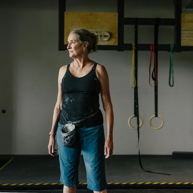 Older woman with chalk on her clothing and a chalk bag, stands in an indoor climbing gym with climbing rings and equipment.