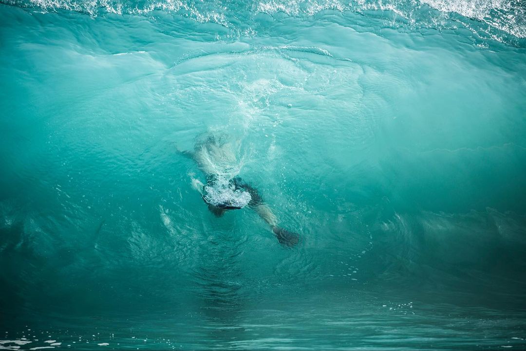 Underwater view of a swimmer beneath a turquoise wave, capturing the dynamic movement and transparency of the water.