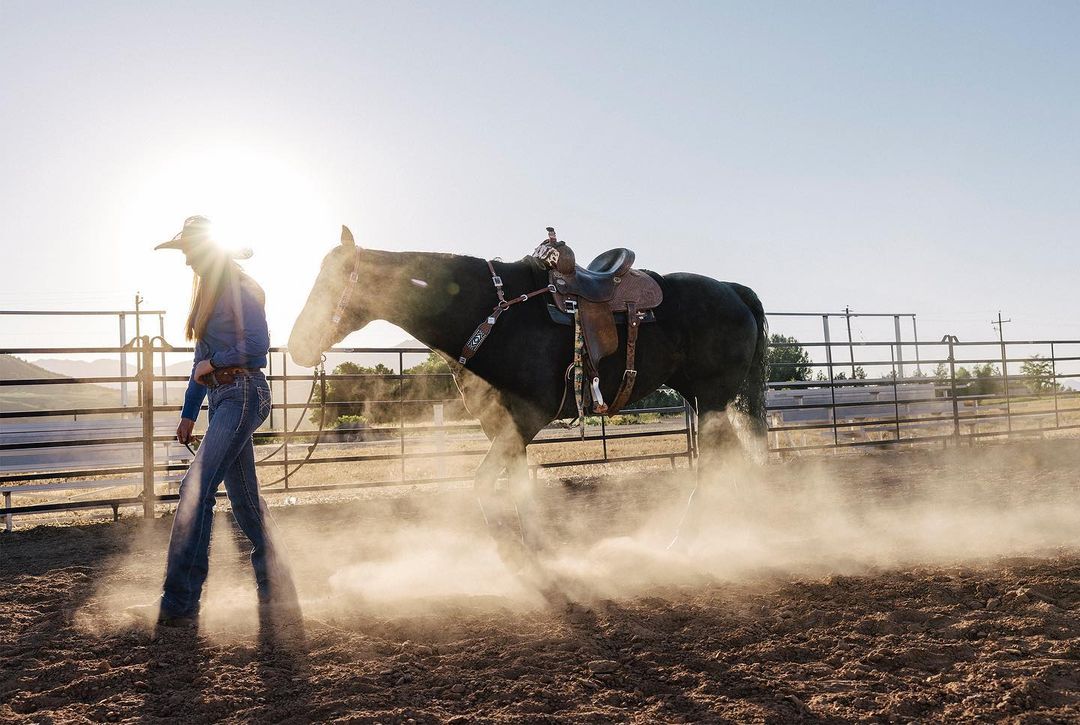 A woman in cowboy attire walks a horse in an outdoor paddock, with dust kicking up in the sunlight, showcasing rural ranch life.