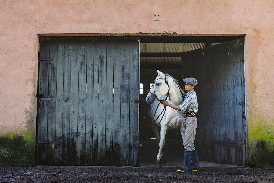 A young man wearing a hat stands next to a white horse in front of the weathered blue doors of a stable in the countryside.