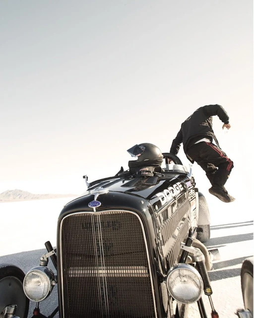 Action shot of a vintage black Ford hot rod with driver mid-air in racing gear on a white, flat landscape under a bright sky.