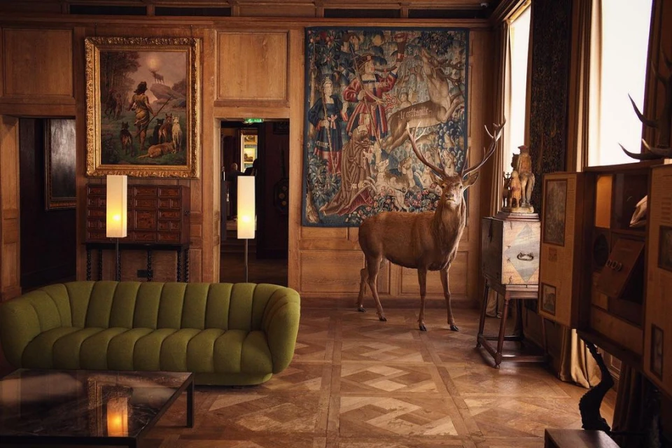 Ornate living room features a mounted stag, olive-green sofa, art, and decorative wood paneling, creating a vintage, high-end interior.