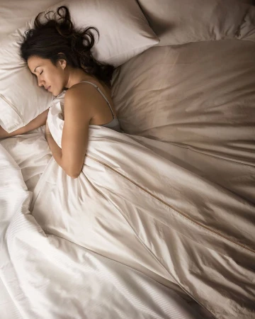 Woman sleeps in bed under white comforter with head resting on pillow. Indoor shot of bedding and comfort in bedroom.