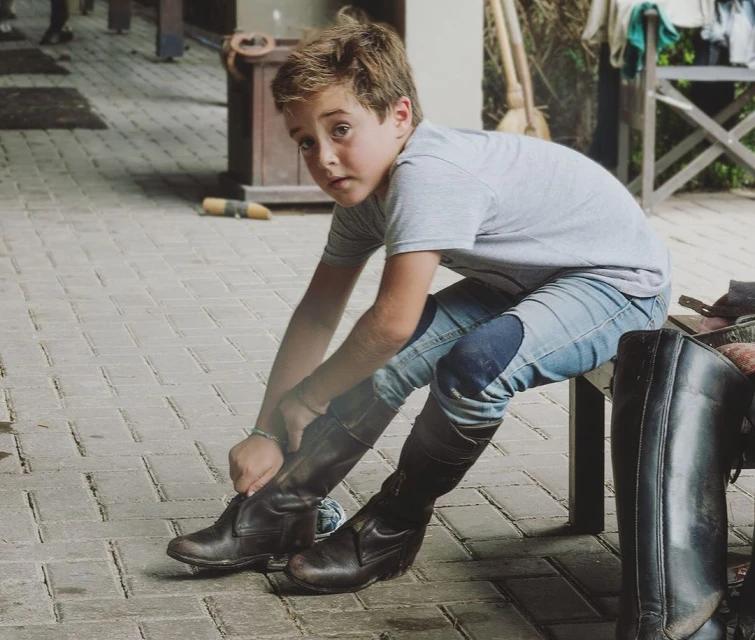Young boy sitting and tying the laces of his riding boots outdoors. He wears jeans, a t-shirt, and looks toward the camera.