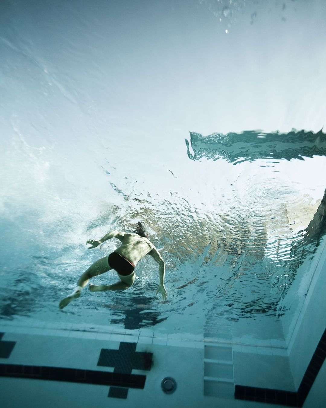 Underwater view of a man swimming laps in an indoor pool with blue tiled walls and clear water, captured with a wide angle lens.