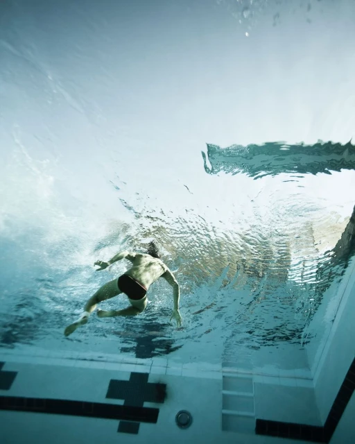 Underwater view of a man swimming laps in an indoor pool with blue tiled walls and clear water, captured with a wide angle lens.