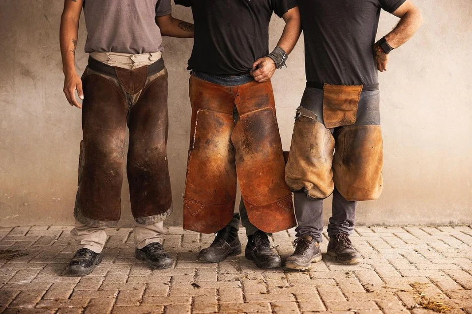 Three adult men stand in work clothing that includes protective leather chaps, posed in front of a neutral wall and brick ground.
