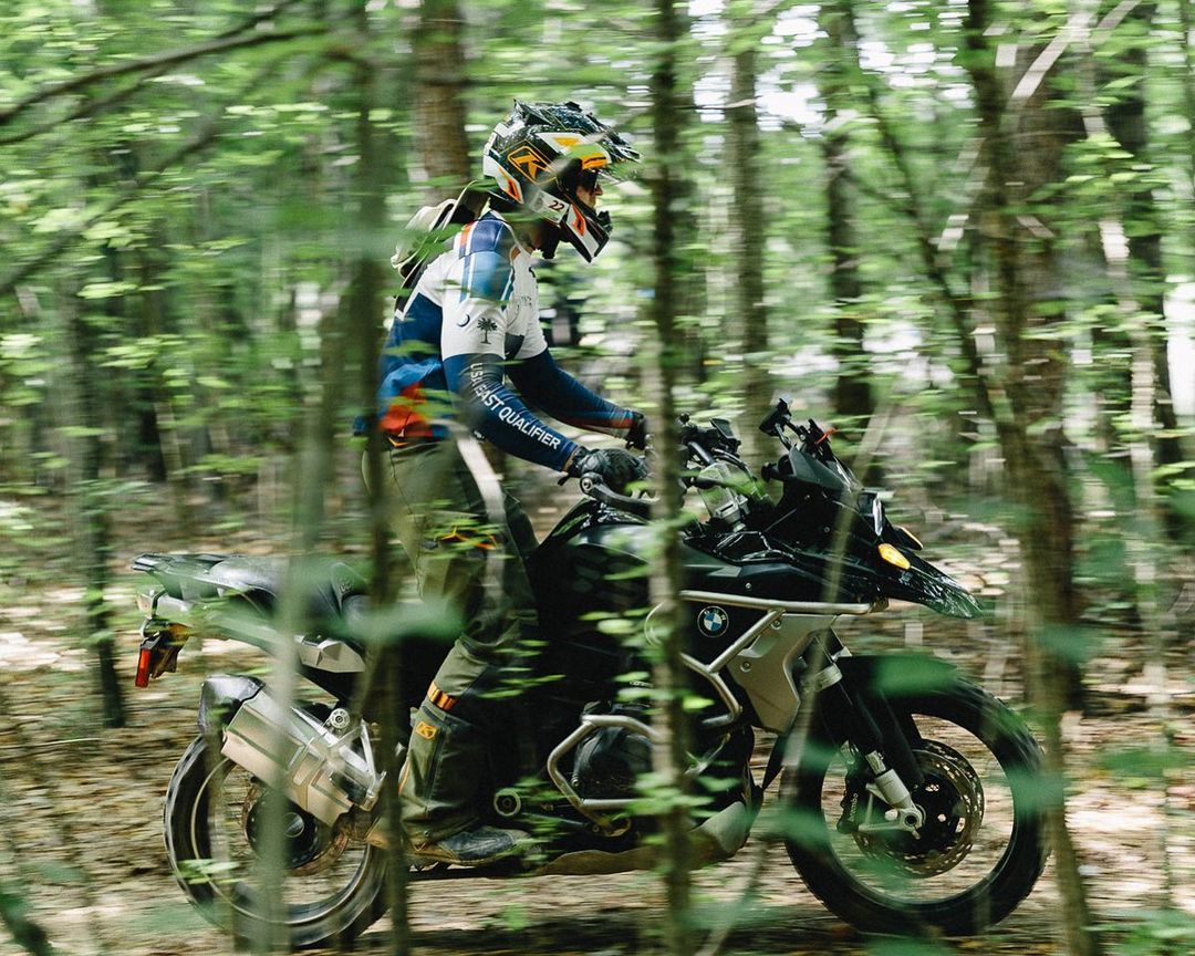 Person riding a BMW motorcycle on a dirt road through a sunlit forest, wearing a helmet and protective gear, trees blurring in the background.