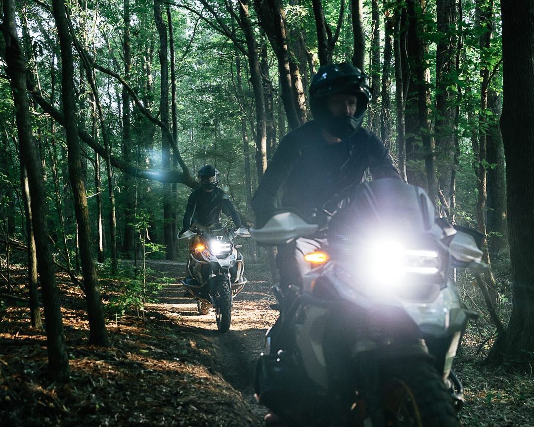 Two people riding adventure motorcycles on a dirt path through a dense, green forest with bright headlights illuminating the path.
