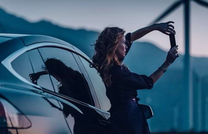 Adult woman takes a cell phone picture while leaning against the side of a dark coloured car with mountains in the background at dusk.
