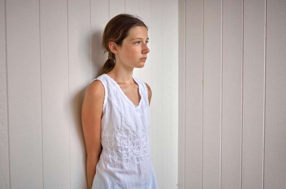 Young woman in white dress standing against a white wall. Medium shot of a woman looking left with fair skin and light brown hair.