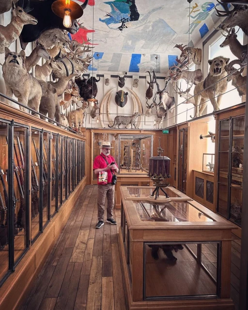 Man stands in a taxidermy-filled hall with animal heads, trophy mounts, and glass display cases containing rifles, creating a museum-like setting.